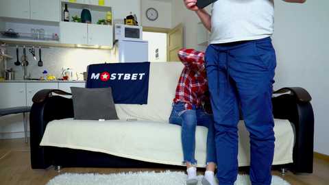 Media: A video depicts a young woman in a red plaid hoodie and jeans sitting on a white couch in a modern kitchen. A man in blue work pants and a white shirt stands beside her.