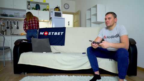 Media: Video of a man in a gray T-shirt and blue pants watching TV on a white couch in a modern, minimalist kitchen.