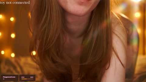 Media: Video of a fair-skinned woman with long brown hair, wearing a top, leaning forward. Background features blurred, warm-toned lights and a cozy, intimate setting.