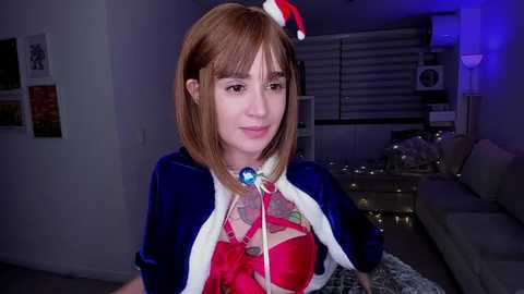 Media: A video of a young woman with straight, light brown hair and fair skin wearing a red bra, a blue velvet jacket, and a Santa hat, indoors with Christmas decorations and a gray couch in the background.