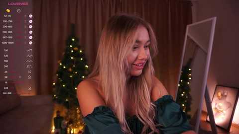 Media: Video of a smiling, blonde woman in a green off-shoulder dress, looking at a mirror in a cozy, dimly lit room with Christmas decor and warm lighting.