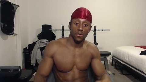 Media: Video of a shirtless, muscular Black man with a red baseball cap, sitting in a gym with weights and exercise equipment in the background.