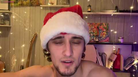 Media: Video of a shirtless young man with a red Santa hat, closed eyes, and a relaxed expression. Background features string lights, framed art, a guitar, and a colorful painting.