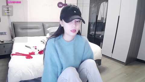 Media: A video of an East Asian woman with long black hair, wearing a black cap, light blue sweater, and gray pants, sitting on a bed in a modern, minimalist bedroom.