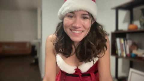 Media: Video of a smiling woman with wavy dark hair wearing a red Santa hat and dress, standing in a modern living room with shelves and a brown couch.