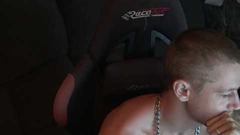 Media: Video of a shirtless, fair-skinned man with a short buzz cut, wearing a silver chain necklace, lying on a dark, reclining car seat, eyes closed, in a dimly lit car interior.