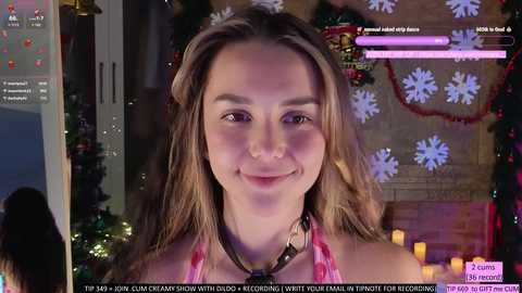 Media: Video of a young woman with long brown hair, smiling, in a festive indoor setting with holiday decorations. She wears a pink top and a black choker. The background includes a fireplace, snowflakes, and candles. Text overlays the image with a gaming context.