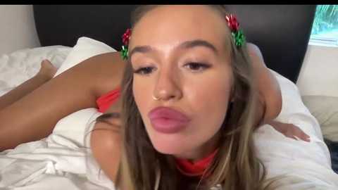 Media: Video of a young woman with fair skin, long brown hair, and red lipstick, lying on a white bed, wearing a red top, with a festive headband.