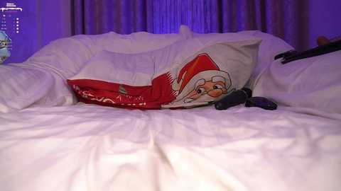 Media: Video of a messy white bed with a red Santa pillow and a black remote control, set against dark curtains.