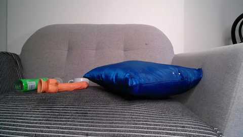 Media: Video of a grey, textured sofa with a blue pillow and an orange and green drink bottle on a striped cushion. Minimalistic, indoor setting.