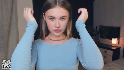 Media: Video of a young woman with light skin, long straight blonde hair, wearing a light blue long-sleeve top, flexing her arms, indoors with a TV, lamp, and beige curtain in the background.