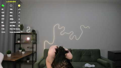 Media: Video of a woman in a green dress sitting on a green couch, holding a black cat, with a neon light drawing a wavy line on a gray wall.