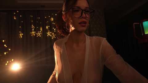 Media: Video of a woman with dark hair, glasses, and a sheer top, in a dimly lit room with golden string lights and a TV in the background.