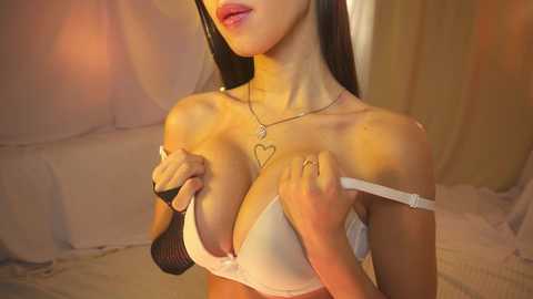 Media: Video of a young woman with long black hair, wearing a white bra, heart necklace, and black mesh glove, posing seductively in a dimly lit bedroom.