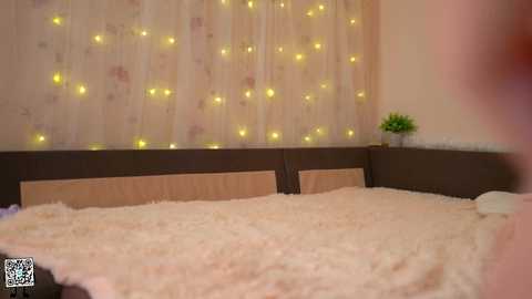 Media: A video of a cozy bedroom with a bed covered in a fluffy white blanket, a wooden headboard, and a beige floral wallpaper adorned with warm yellow fairy lights.