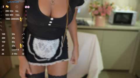 Media: Video of a woman wearing a revealing black and white French maid outfit, featuring a low-cut top and lace apron, standing in a kitchen with a microwave and flowers in the background.