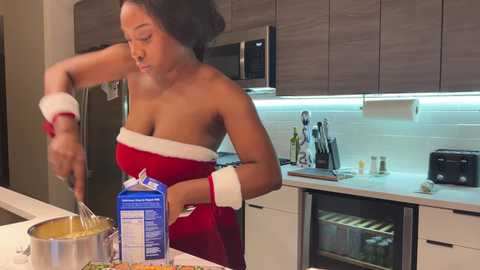 Media: Video of a Black woman with medium skin tone and medium-length hair, wearing a red strapless dress with white fur trim, mixing a cake batter in a modern kitchen with stainless steel appliances.