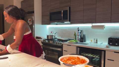 Media: Video of a young African American woman in a red apron cooking in a modern kitchen with white cabinets, stainless steel appliances, and a white tiled backsplash.