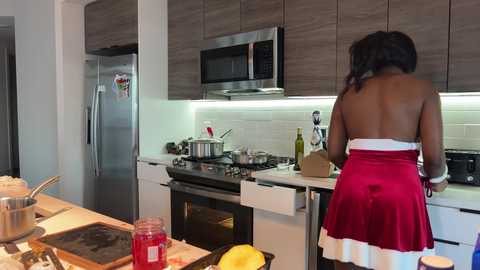 Media: Video of a modern kitchen with a woman in a red dress and white apron cooking at the stove. Stainless steel appliances, white subway tiles, and a wooden island with cooking utensils are visible.