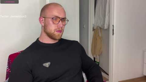 Media: Video of a bald, bearded white man with glasses, wearing a black long-sleeve shirt, seated indoors. Background includes a white door, a red chair, and a hanging white towel.