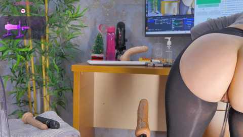 Media: A video of a woman in a black thong, bent over a desk, showing her bare buttocks. The room has a modern, colorful decor with a large screen displaying text.