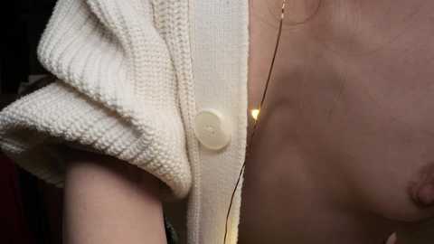 Media: Video of a fair-skinned woman with a soft, textured white cardigan and a thin gold necklace, partially exposed breasts visible.