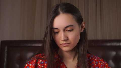 Media: Video of a young woman with long dark hair, wearing a red floral-patterned top, seated on a dark leather couch, gazing downward in a contemplative manner. Background features beige curtains.