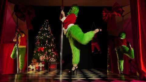 Media: A vibrant video of a green-skinned woman in a red Santa suit and green fur pants, dancing on a pole, surrounded by festive Christmas decor and red curtains.