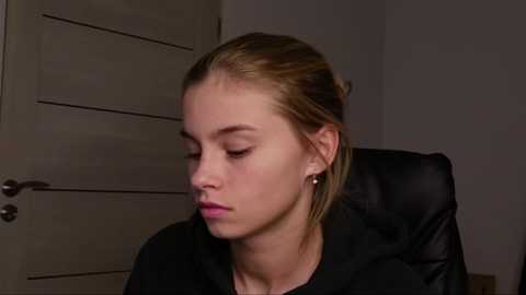 Media: Video of a young Caucasian woman with light skin, light brown hair, and small round earrings, wearing a black shirt, sitting indoors with a neutral expression.