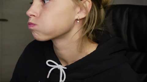 Media: Video of a young woman with light skin and blonde hair, wearing a black hoodie with a white drawstring, looking contemplative and slightly pensive.