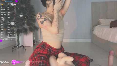 Media: A video shows a slender, tattooed woman in a beige crop top and red plaid pants, kneeling in a bedroom. A Christmas tree and bed are in the background.