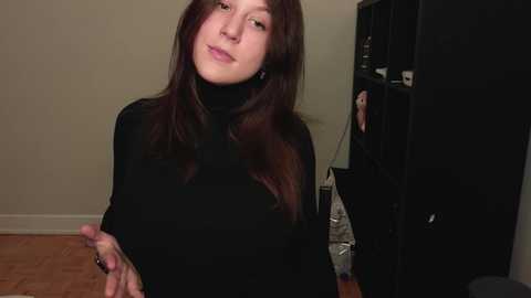 Media: A video of a young woman with straight, shoulder-length brown hair, wearing a black turtleneck, standing indoors with a neutral expression. Background includes a black bookshelf and a white wall.