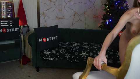 Video of a naked man with a beard, holding a Christmas ornament, standing in a room with a green couch, a black and white patterned blanket, and a decorated Christmas tree.