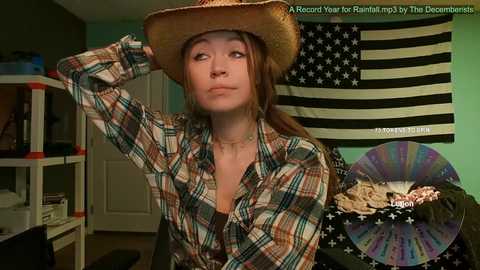 Media: Video of a young woman with auburn hair wearing a cowboy hat, plaid shirt, and a neutral expression, standing in a room with a U.S. flag and colorful decorations.