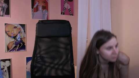 Media: Video of a young woman with long brown hair, wearing a brown top, standing in a room with a black massage chair and colorful posters on a pink wall.