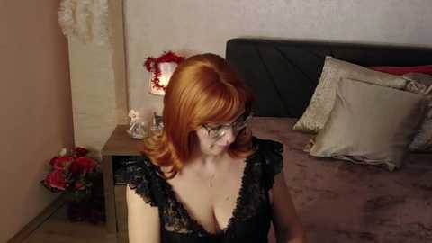 Media: Video of a fair-skinned, red-haired woman with glasses, wearing a black lace dress, sitting on a bed with pink sheets, surrounded by pillows, in a cozy, dimly lit bedroom.