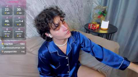 Media: Video of a young man with curly dark hair, wearing a blue satin robe, sitting on a beige couch. Background includes a black table with a white candle and red flowers, and a gray curtain.