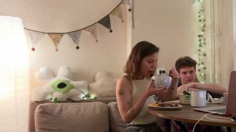 Media: Video of a young woman with shoulder-length brown hair in a beige tank top, holding a spoon near a wooden table, with a young man sitting across from her, both looking at a laptop, in a cozy living room with pastel decor.