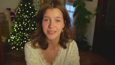 Media: A video of a smiling young woman with wavy brown hair, wearing a light-colored blouse, in a warmly lit room with a Christmas tree and potted plants in the background.