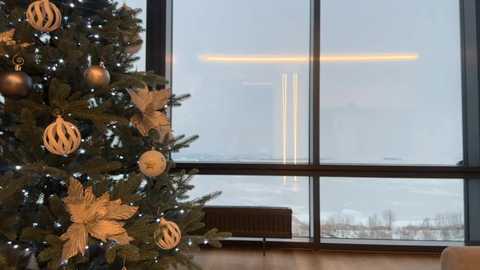 Media: Video of a modern living room with a decorated Christmas tree on the left, adorned with golden ornaments, and a large window showing a snowy landscape outside.