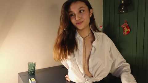 Media: Video of a young woman with long, straight brown hair, light brown skin, and a slender build, wearing a white blouse unbuttoned to reveal her chest, standing in a dimly lit room with beige walls, green door, and a teal glass on a table.