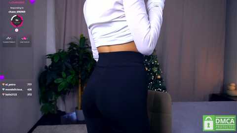 Media: Video of a woman in a white long-sleeved top and high-waisted black pants, standing indoors near a Christmas tree. Social media icons and text overlay.