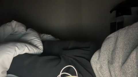 Media: A dimly lit video of a person in bed, wearing a black shirt with a white emblem. The bedding is messy with white pillows and gray blanket. The background is dark and indistinct, creating a moody atmosphere.