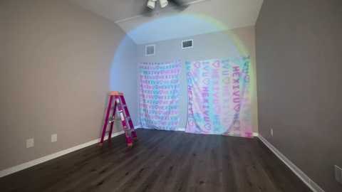 Media: Video of a room with beige walls, dark wooden floor, and a pink ladder. Rainbow light shines on a colorful, patterned curtain. Ceiling fan.