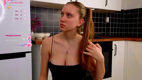 Media: Video of a slender, fair-skinned woman with long brown hair tied in a ponytail, wearing a black spaghetti-strap top, standing in a modern kitchen with white cabinets, black tiled backsplash, and a fridge displaying \"The Love Diet\" information.