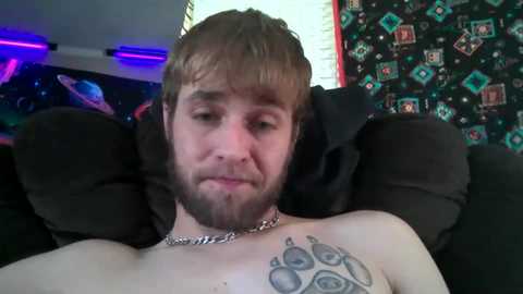 Media: A close-up video of a bearded man with short, light brown hair, wearing a silver chain necklace and a tattoo of a paw print on his right shoulder. Background features a dark couch and a wall with colorful, cartoon-themed wallpaper.