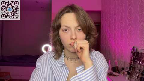 Video of a young woman with light skin and shoulder-length brown hair, wearing a striped shirt and necklace, with a pensive expression, sitting in a dimly lit room with pink lighting.