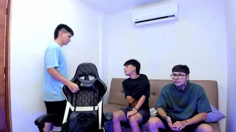 Media: Video of three young men in a simple room with white walls, an air conditioner, and a beige couch. One man stands, adjusting a black gaming chair; two others sit, one in a green shirt, the other in black.