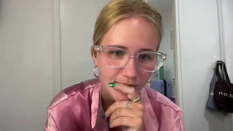 Media: A video of a blonde woman with fair skin, wearing clear glasses, a pink satin robe, and a green nail polish, holding a toothbrush in a bathroom with a white door and a brown bag.