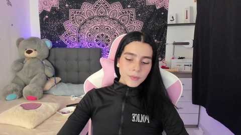 Media: Video of a young woman with long black hair, wearing a black zip-up jacket, seated in a pink gaming chair, in a cozy room with a gray teddy bear, a black and white mandala tapestry, and a desk with personal items.